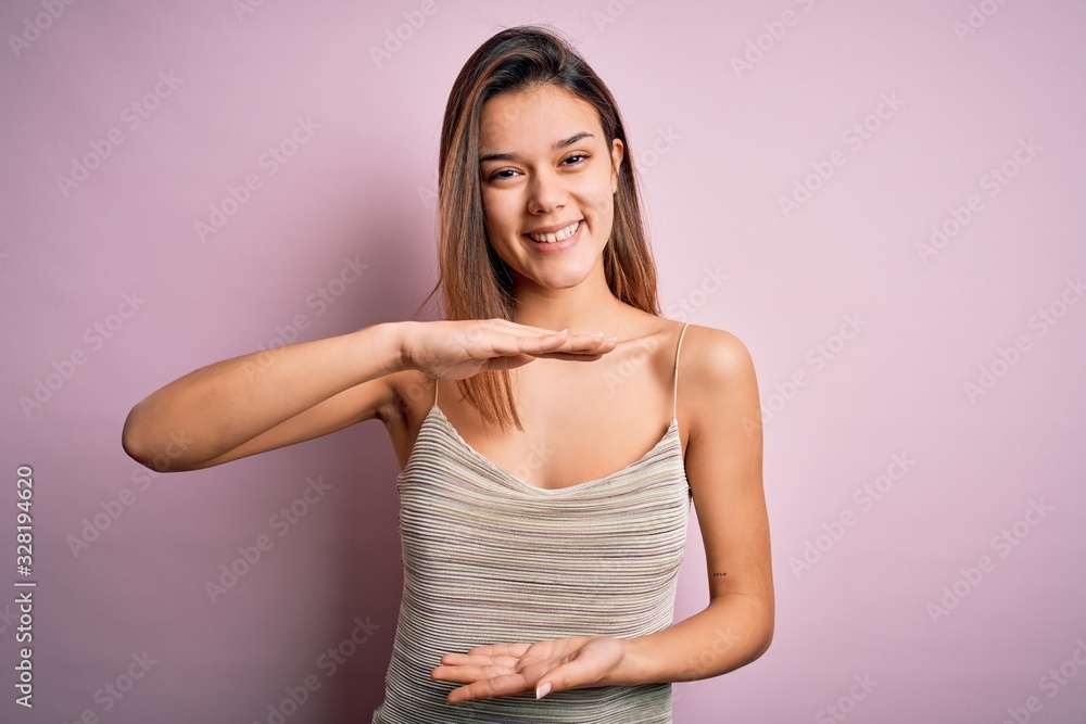 Young beautiful brunette girl wearing casual striped t-shirt over isolated pink background gesturing with hands showing big and large size sign, measure symbol. Smiling looking at the camera. Measure Young beautiful brunette girl wearing casual striped t-shirt over isolated pink background gesturing with hands showing big and large size sign, measure symbol. Smiling looking at the camera. Measure