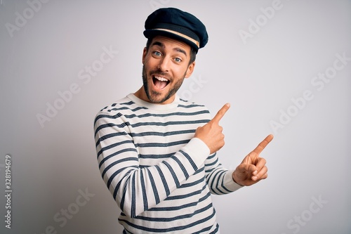 Young handsome sailor man with beard wearing navy striped uniform and captain hat smiling and looking at the camera pointing with two hands and fingers to the side. photo