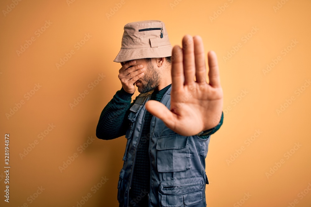 Handsome tourist man with beard on vacation wearing explorer hat over yellow background covering eyes with hands and doing stop gesture with sad and fear expression. Embarrassed and negative concept. Handsome tourist man with beard on vacation wearing explorer hat over yellow background covering eyes with hands and doing stop gesture with sad and fear expression. Embarrassed and negative concept.