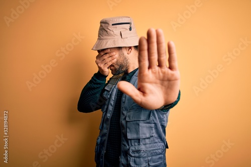 Handsome tourist man with beard on vacation wearing explorer hat over yellow background covering eyes with hands and doing stop gesture with sad and fear expression. Embarrassed and negative concept.