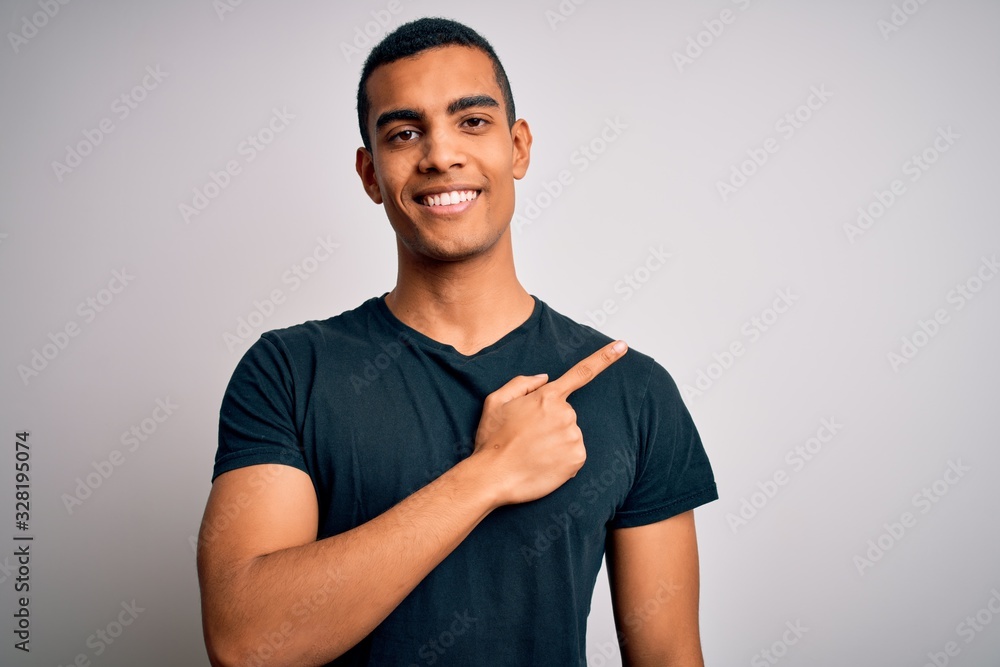 Young handsome african american man wearing casual t-shirt standing over white background cheerful with a smile on face pointing with hand and finger up to the side with happy and natural expression Young handsome african american man wearing casual t-shirt standing over white background cheerful with a smile on face pointing with hand and finger up to the side with happy and natural expression