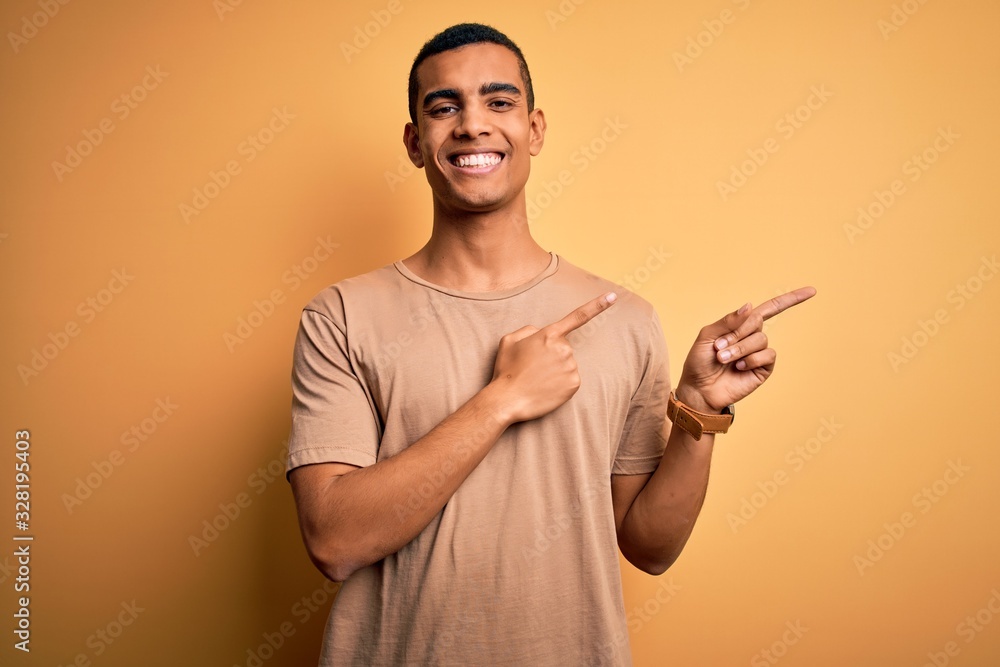 Young handsome african american man wearing casual t-shirt standing over yellow background smiling and looking at the camera pointing with two hands and fingers to the side. Young handsome african american man wearing casual t-shirt standing over yellow background smiling and looking at the camera pointing with two hands and fingers to the side.
