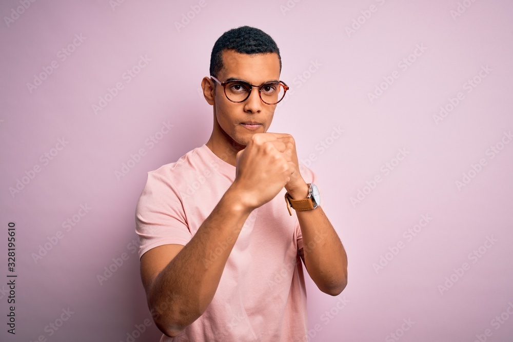 Handsome african american man wearing casual t-shirt and glasses over pink background Ready to fight with fist defense gesture, angry and upset face, afraid of problem Handsome african american man wearing casual t-shirt and glasses over pink background Ready to fight with fist defense gesture, angry and upset face, afraid of problem