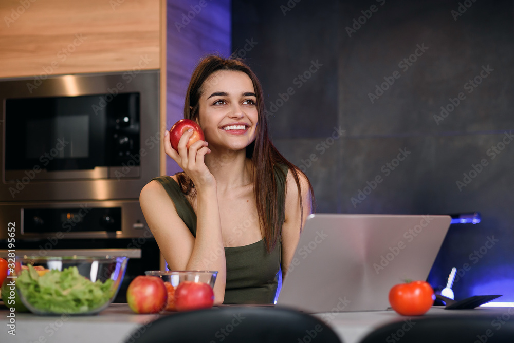 Image of a beautiful young student girl with apple sitting indoors using laptop computer.