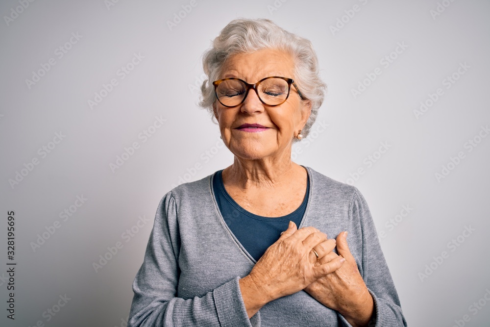 Senior beautiful grey-haired woman wearing casual sweater and glasses over white background smiling with hands on chest with closed eyes and grateful gesture on face. Health concept. - obrazy, fototapety, plakaty 