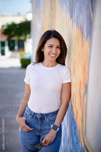 Smiling woman in white T-shirt and blue jeans standing on the street next to wall holding sunglasses