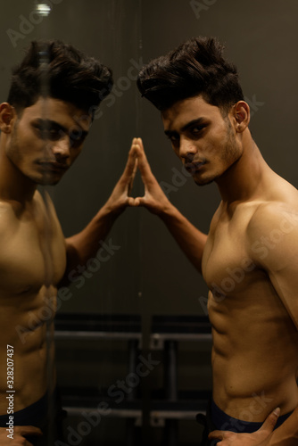 An young and handsome Indian Bengali brunette man and his mirror image with muscular body doing exercise in a multi gym. Fitness and Indian lifestyle.