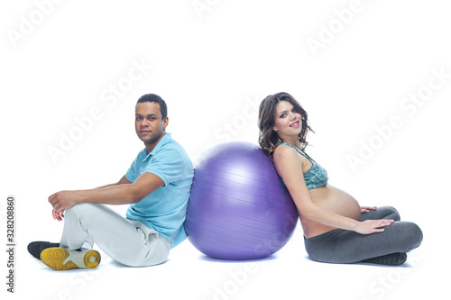 A young couple and their wife in the last months of pregnancy are sitting on a sports mat near the ball. Black man and white woman. The concept of a happy couple of different races preparing together
