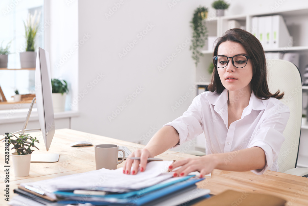 Attractive young businesswoman sitting at desk in office and finding documents in stack Attractive young businesswoman sitting at desk in office and finding documents in stack