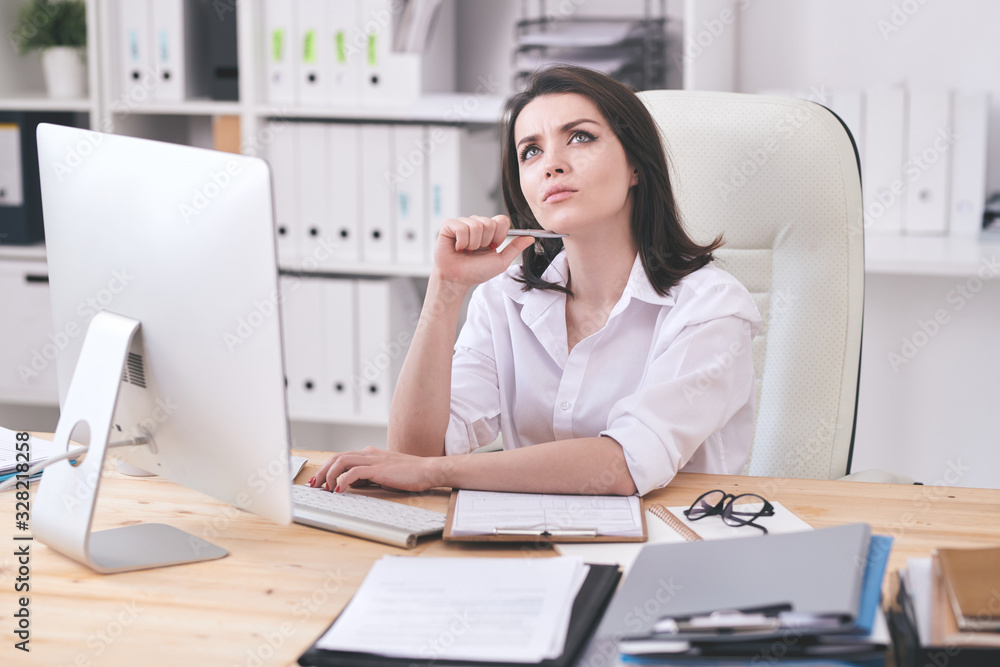 Pensive young business lady in white blouse sitting at table in office and thinking about online project Pensive young business lady in white blouse sitting at table in office and thinking about online project