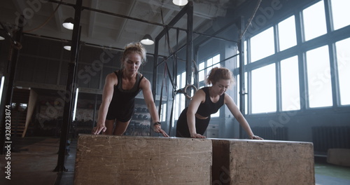 Fotografi Slow motion two young beautiful female athletes working out, doing push-ups and clapping together in atmospheric gym