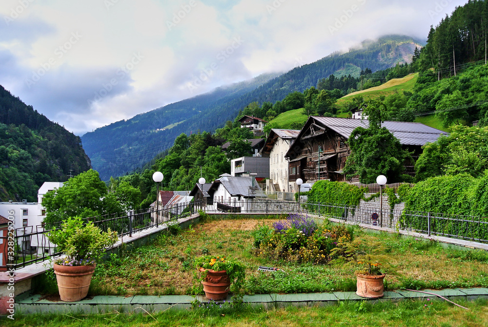 Landeck, Austria - Houses in a small town, amid green alpine mountains covered with forest, flower pots in the foreground, gray clouds in the sky, in the summer afternoon.
