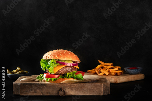 Burger with beef, cheese, onion, tomato, and green salad, a side view on a dark background with French fries and pickles, with a place for text