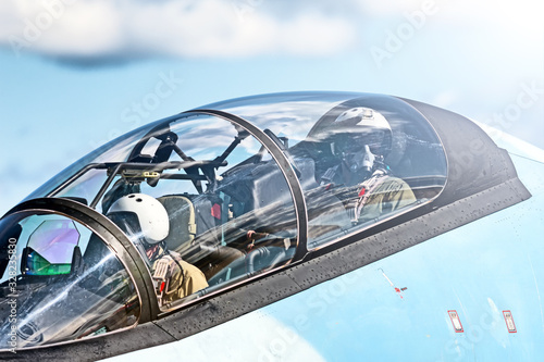 military pilot in fighter jet cockpit waving hand closeup against blue sky background. Fighter jet crew on duty in combat aircraft Detailed view of military airplane glass canopy © vaalaa