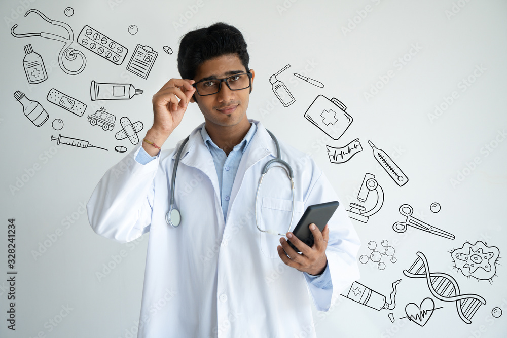 Indian doctor adjusting glasses with hand drawn medical sketches. Serious experienced young clinical worker in lab coat checking sms and looking at camera. Online medicine concept Indian doctor adjusting glasses with hand drawn medical sketches. Serious experienced young clinical worker in lab coat checking sms and looking at camera. Online medicine concept