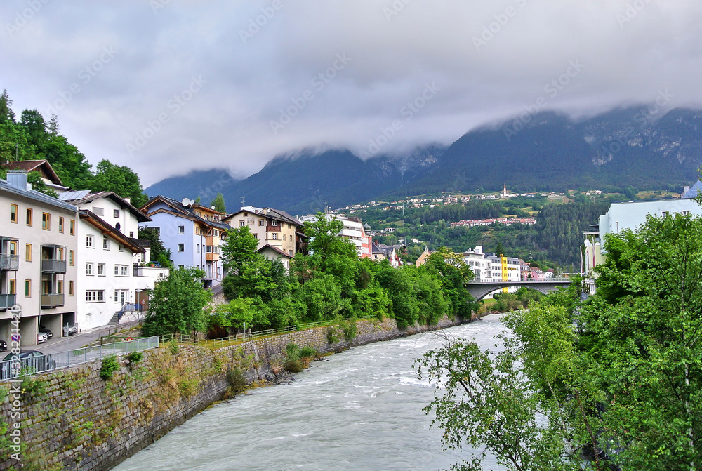 Landeck, Austria - Houses in a small town on the banks of a mountain river among green trees, amid alpine mountains covered with clouds, in the summer afternoon.