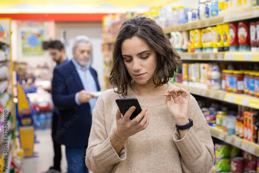 Woman using cell phone in supermarket. Young woman reading checklist via smartphone and choosing products in grocery store. Supermarket concept Woman using cell phone in supermarket. Young woman reading checklist via smartphone and choosing products in grocery store. Supermarket concept