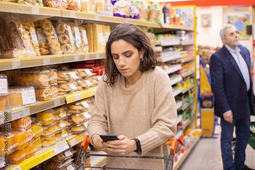 Woman using smartphone in grocery store. Young woman reading checklist via smartphone and choosing goods in grocery store. Supermarket concept