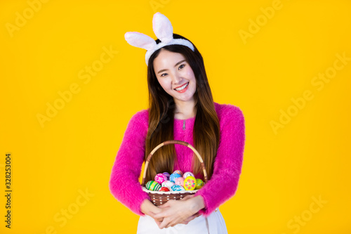 Easter holiday concept, Happy Asian Young woman wearing bunny ears hand holding a basket with colorful Easter eggs In isolated on Yellow blank copy space studio background.