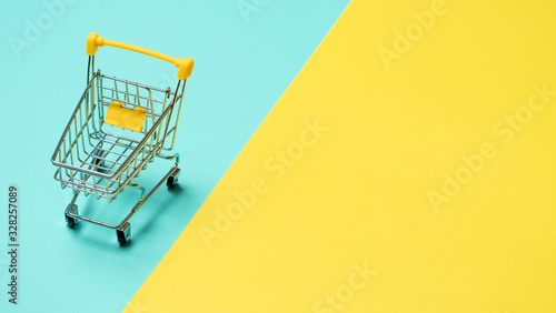 Empty miniature shopping cart on blue and yellow background. Toy trolley on bright colorful background, copy space for text or design.