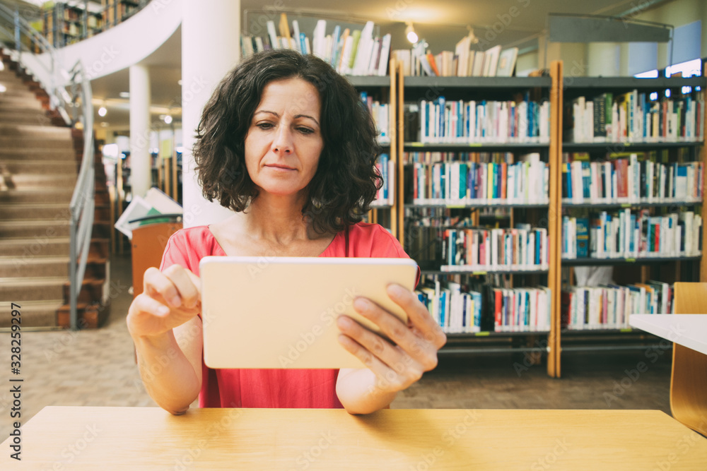 Focused female gadget user watching video content in public library. Middle aged Caucasian woman sitting at desk, using tablet, looking at screen. Video content concept Focused female gadget user watching video content in public library. Middle aged Caucasian woman sitting at desk, using tablet, looking at screen. Video content concept