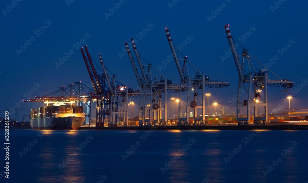 A long exposure in the port of Hamburg at night. A container ship lies at the container terminal with many cranes. The lights reflect in the water. Many containers are stacked on land.