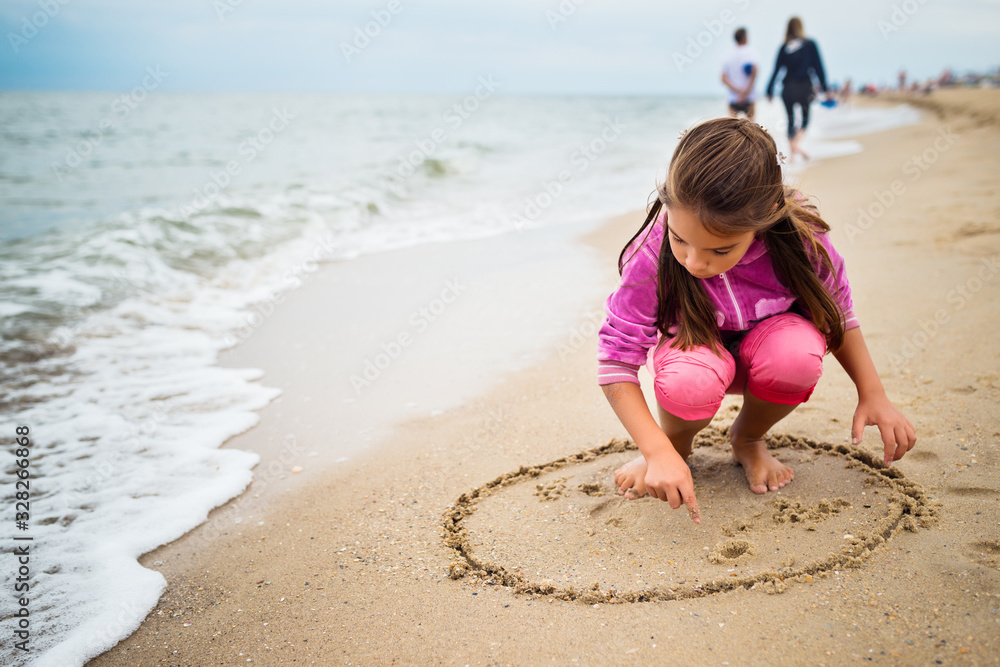 Unidentified tanned little girl draws on the sand near the stormy sea waves on a sunny warm summer day. The concept of the long-awaited summer vacation at sea Unidentified tanned little girl draws on the sand near the stormy sea waves on a sunny warm summer day. The concept of the long-awaited summer vacation at sea