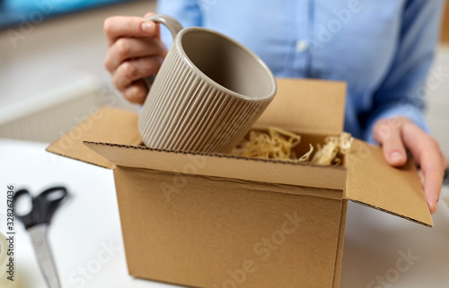 delivery, mail service, people and shipment concept - close up of woman packing mug to parcel box with straw filler at post office