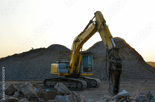 Large tracked excavator with hydraulic hammer breaks asphalt at a construction site on the background sunset. Breaking rock and reinforced Concrete. Road repair, asphalt replacement. Demolition tools