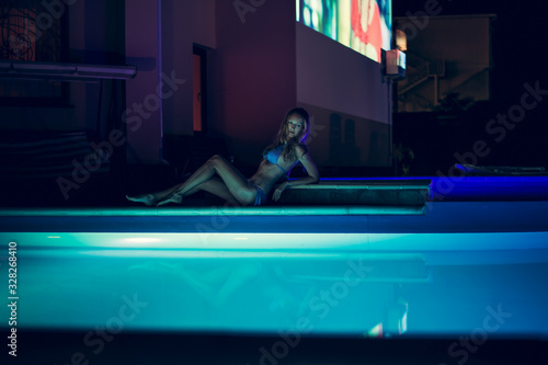 Young slim girl in swimsuit sits at night on the edge of the pool with backlight