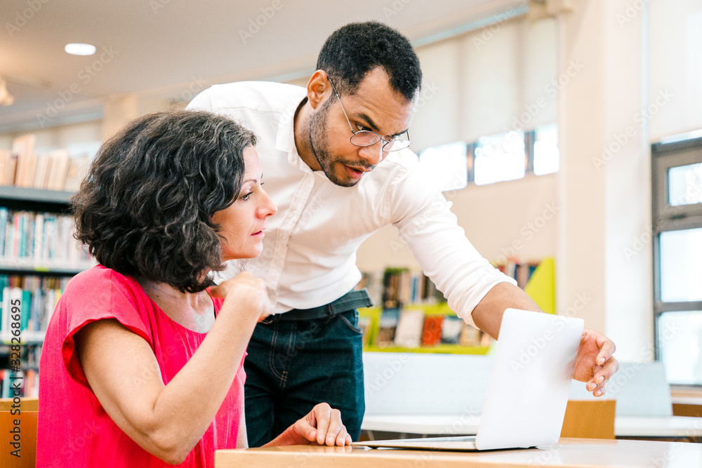 Instructor checking student work in library. Man and woman in casual sitting and standing at desk, using laptop, looking at monitor and talking. Training or course concept