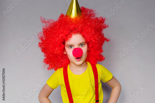 1 April Fool's day concept. Let's celebrate! Funny kid clown playing at home. Girl child with a red wig and a clown's nose is holding a piece of paper for your entry. Copyspace.