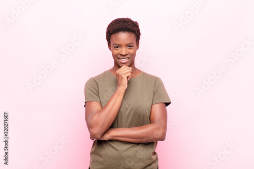 young pretty black womanlooking happy and smiling with hand on chin, wondering or asking a question, comparing options against pink wall photo