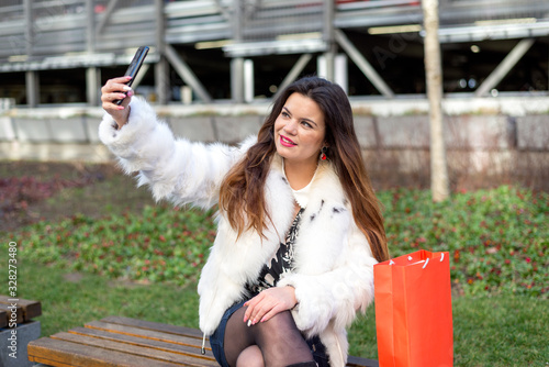 Young woman with shopping bags sitting on the bench and making selfie