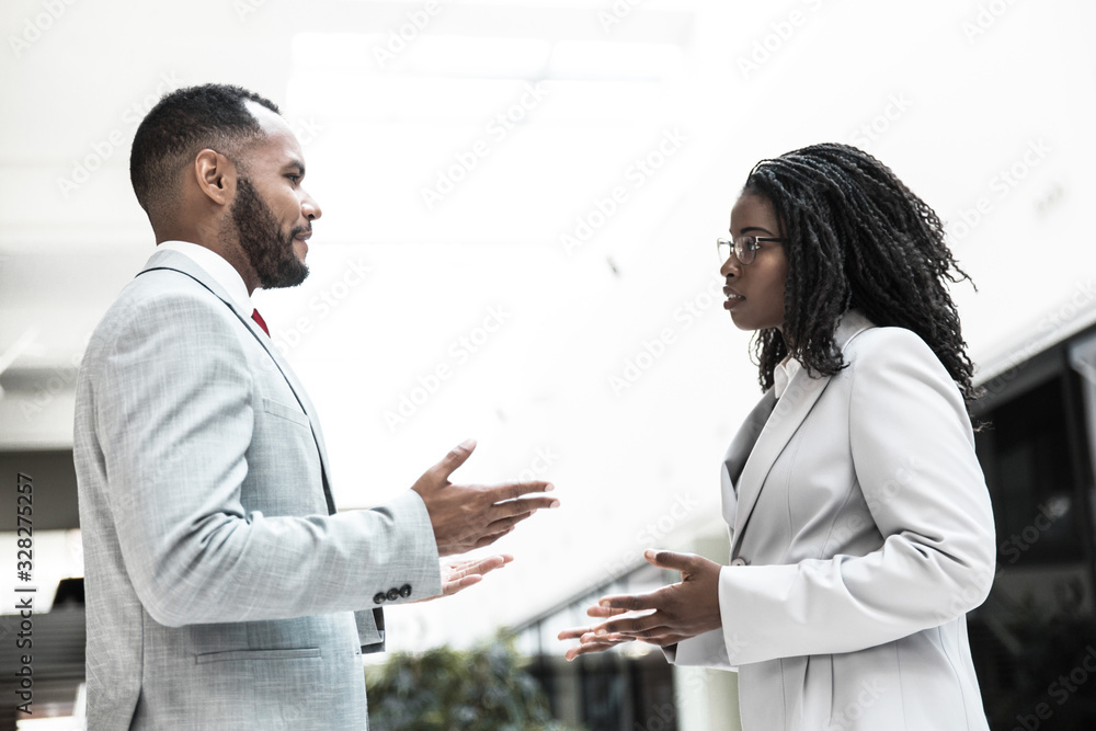 Serious confident business partners discussing project in office hall. Business man and woman standing and talking to each other. Business meeting or negotiation concept Serious confident business partners discussing project in office hall. Business man and woman standing and talking to each other. Business meeting or negotiation concept