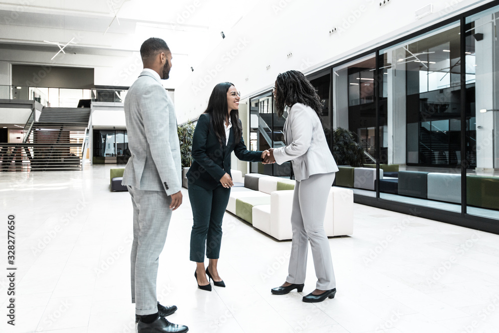 Fototapeta premium Happy successful businesswoman welcoming partners in hallway. Business man and women standing in office hall, shaking hands with each other, talking and smiling. Business relationship concept