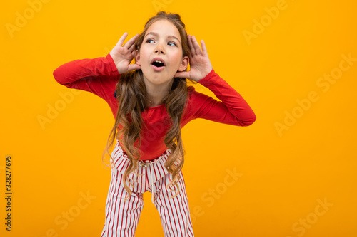 teenager girl with curly hair in a red blouse and striped trousers listens to the sounds while holding hands near the ears on a yellow background with copy space