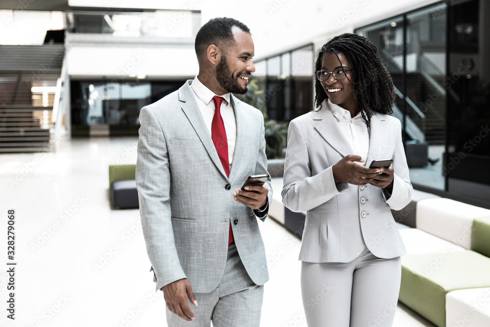 Happy business colleagues using mobile phones and chatting. Business man and woman walking through office hallway, holding smartphones and talking. Corporate communication concept