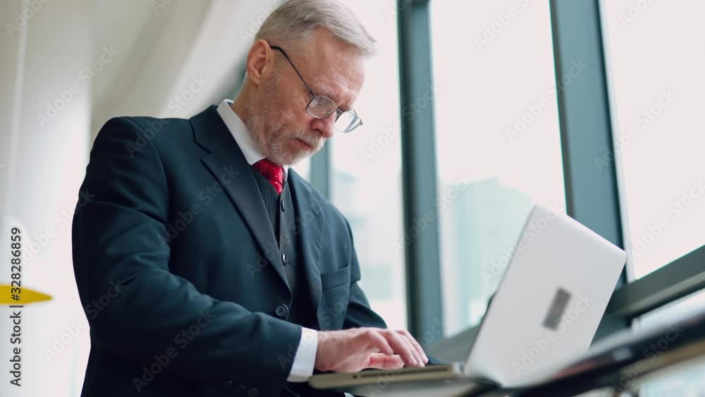 Senior serious businessman standing near window with panoramic city view. Laptop in hands, man thinking while checking e-mails standing near window. Video from the side. Senior serious businessman standing near window with panoramic city view. Laptop in hands, man thinking while checking e-mails standing near window. Video from the side.