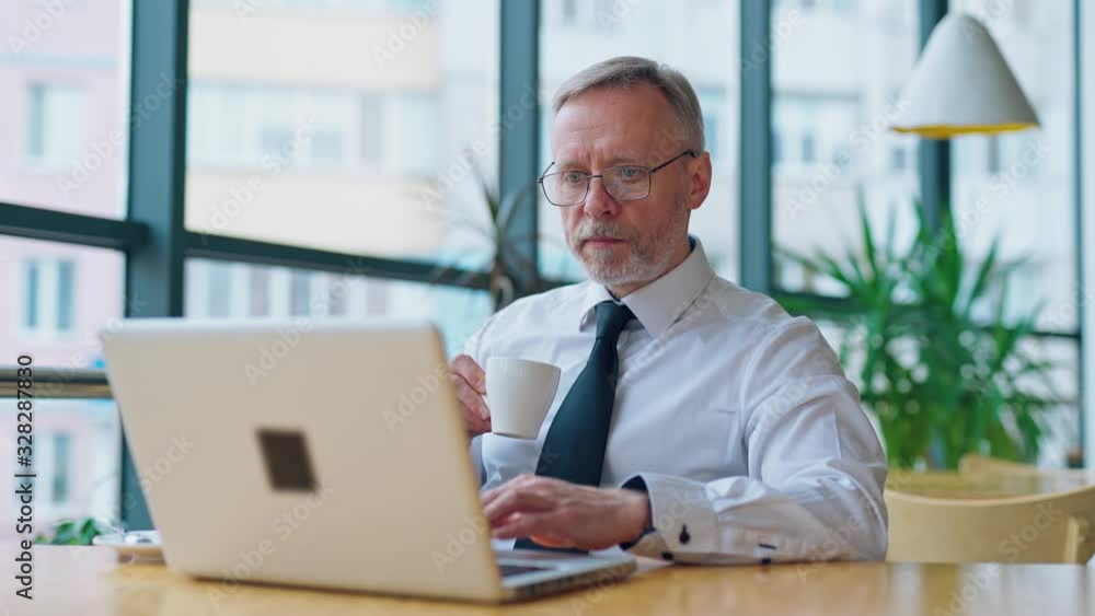 Elderly man is sitting in a light room with big glass windows near computer with a cup of coffee. Working on a latop in modern restaurant during coffee brake. Elderly man is sitting in a light room with big glass windows near computer with a cup of coffee. Working on a latop in modern restaurant during coffee brake.