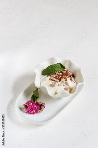 Yogurt with flax and sunflower seeds in a white porcelain bowl and a small pink flower on a white background, with space