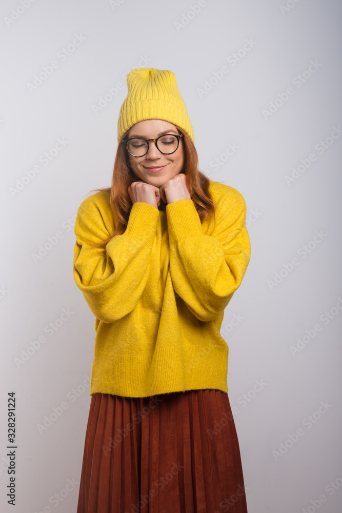 Happy young woman in knitted hat and eyeglasses. Beautiful cheerful young woman in stylish yellow sweater and hat standing with closed eyes isolated on white background. Stylish outfit concept