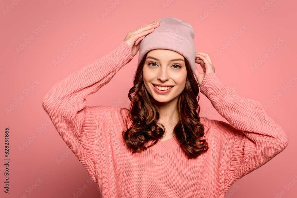 Obraz premium happy girl touching hat while smiling at camera on pink background