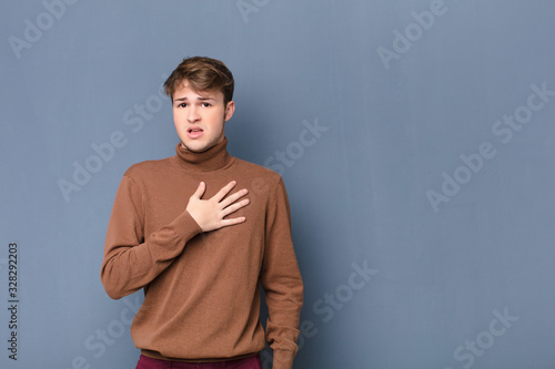 young blonde man feeling shocked, astonished and surprised, with hand on chest and open mouth, saying who, me? isolated against flat wall photo