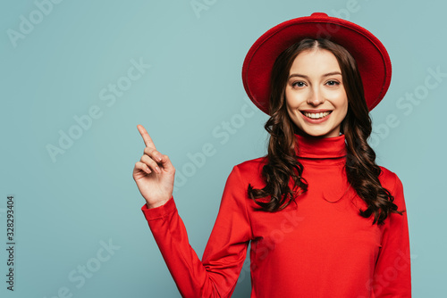 happy stylish girl smiling at camera while pointing with finger isolated on blue