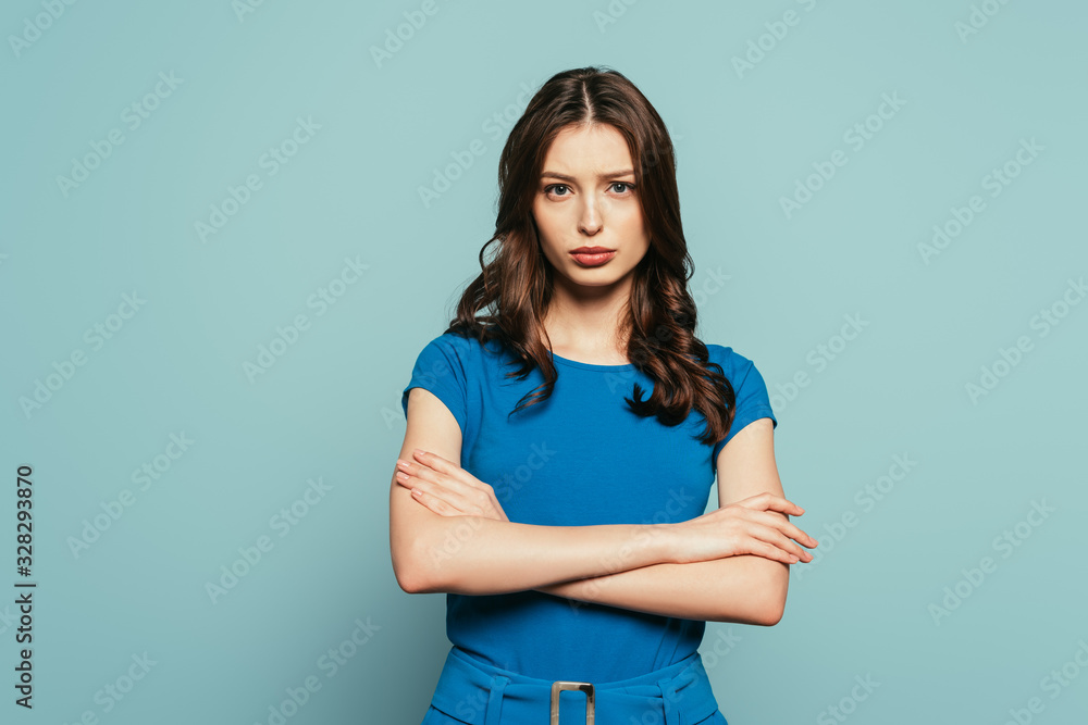 Obraz premium serious, attractive girl looking at camera while standing with crossed arms on blue background