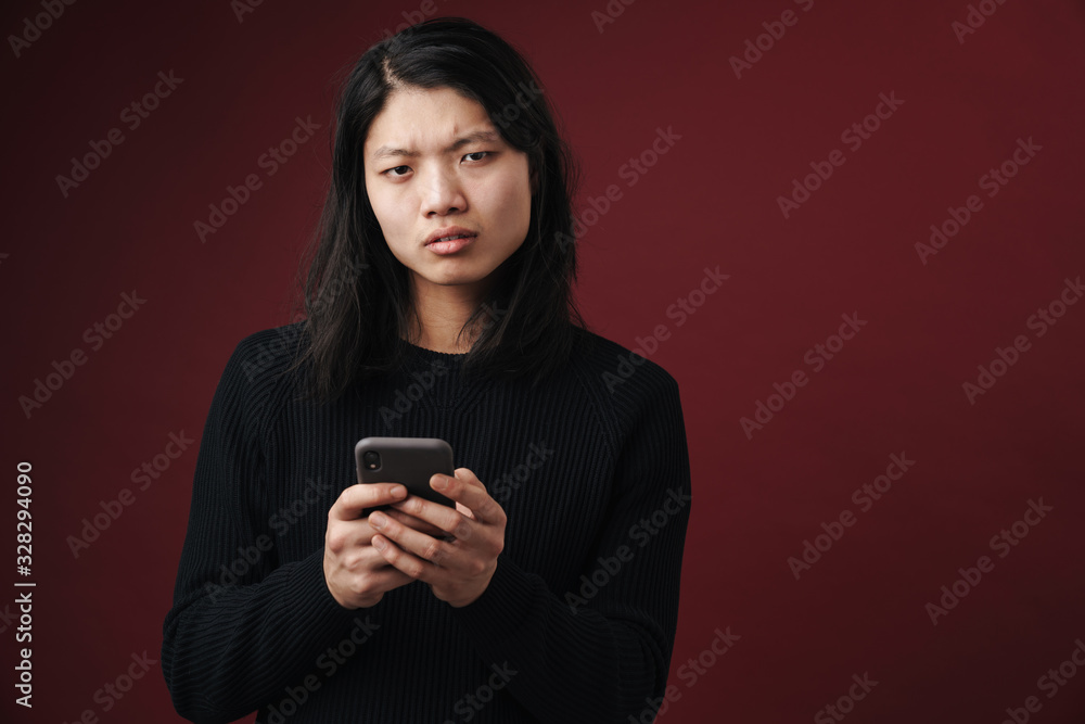 Image of perplexed asian man looking at camera and using cellphone