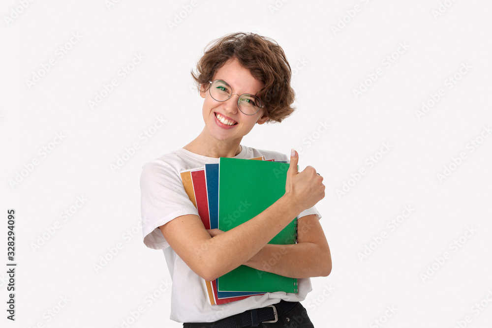 Excited young student in a white shirt and eyeglasses holding colorful folders and showing thumb up Excited young student in a white shirt and eyeglasses holding colorful folders and showing thumb up
