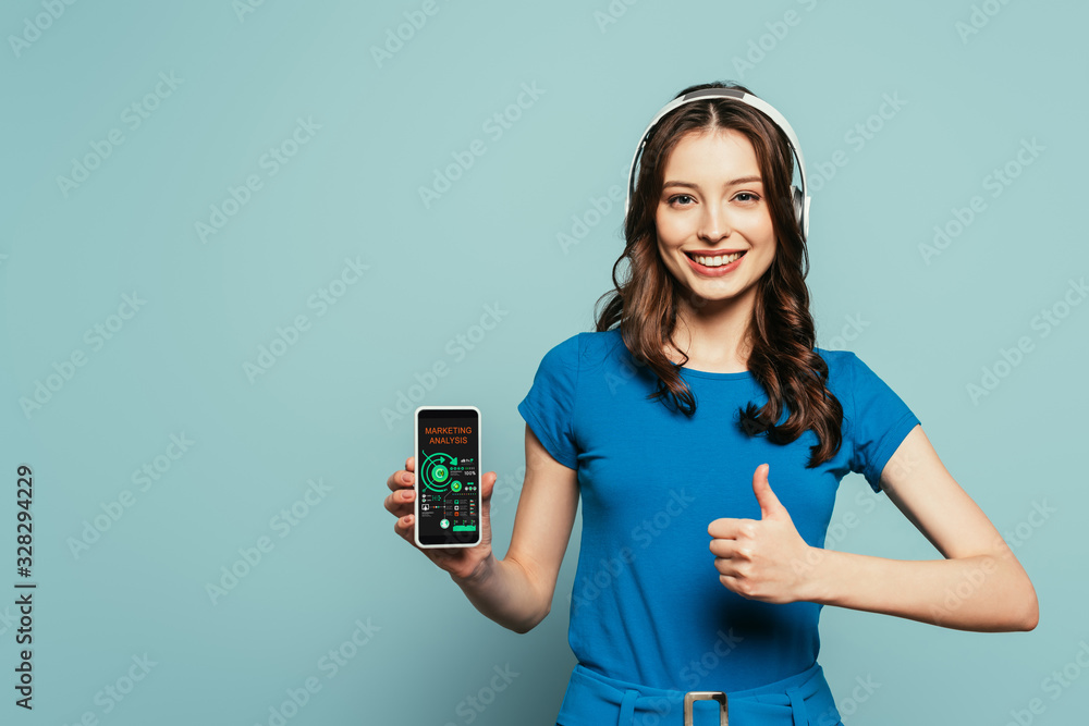 happy girl in wireless headphones showing thumb up while holding smartphone with marketing analysis app on screen on blue background happy girl in wireless headphones showing thumb up while holding smartphone with marketing analysis app on screen on blue background
