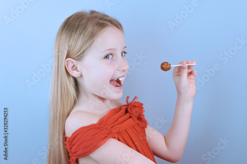 Side view of a beautiful positive little girl joyfully opened her mouth to eat candy on stick on blue background. Sweet tooth kids concept. Nutrition and child health. Copyspace